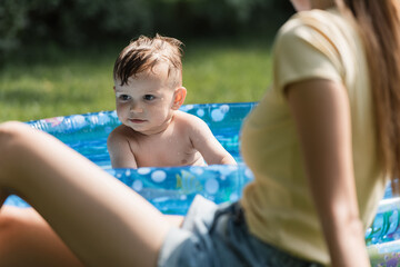 Fototapeta na wymiar toddler boy sitting in inflatable pool near blurred mother