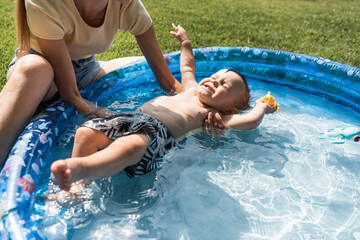 mother supporting toddler son with closed eyes swimming in inflatable pool
