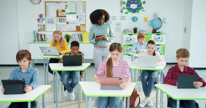 Multi-ethnic diverse junior students studying at school using laptop and tablets, technology, computer science class. African American young female teacher at school teaching bright class of pupils