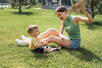 happy young mother holding toy biplane and looking at cheerful toddler son on grass