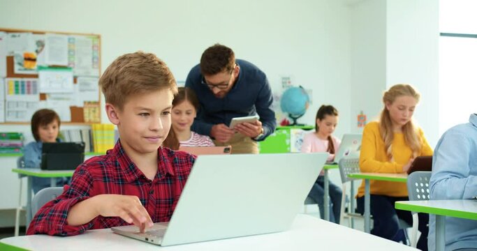 Close up portrait of cute Caucasian teen boy pupil typing on laptop browsing online studying at computer science class, elementary school lesson. Man teacher helping kids using tablet on background