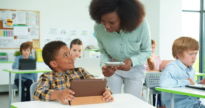 Close up portrait of young beautiful African American woman teacher helping little schoolboy at lesson, elementary school education, computer science class. Junior student tapping on tablet at lesson