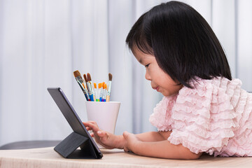 A cute girl is in a good mood choosing her favorite online lessons with a tablet. Asian children smile sweetly. Child enjoy learning online.