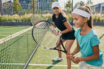 Positive little girl holding a racket while learning to play tennis with her female coach on...
