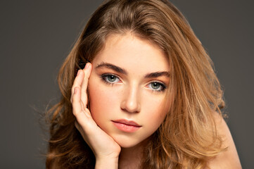 Fototapeta premium Pretty young woman with long brown hair looking at camera. Closeup portrait of an young adult girl with long curly hair. Photo of a fashion model posing at studio. Beauty portrait.
