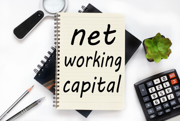 Net Working Capital. an open notebook lies on a closed notebook near a calculator and a magnifying glass on a white workspace.
