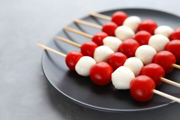 A delicious healthy snack on wooden skewers: balls of mozzarella cheese and red cherry tomatoes on a black plate on a gray background. Selective focus.