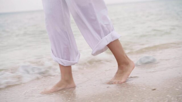 Barefoot Alone Woman Walking Over Sand Sea Beach Close Up Female Feet Goes Along Sea Coast Caucasian Adult Female Dressed White Pants and Shirt Walk on Nature Traveling Concept Low Angle View