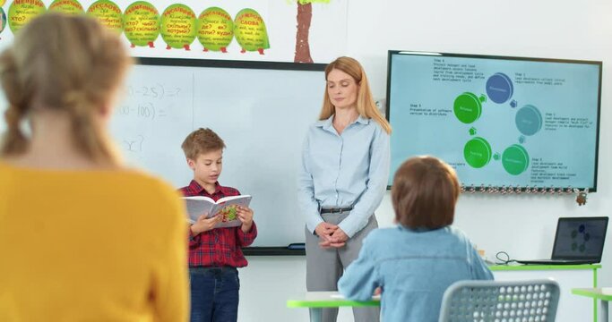 Caucasian pretty young woman teacher standing at school classroom near whiteboard while small junior student boy reading from book in front of classmates. Education, studying, learning, teaching