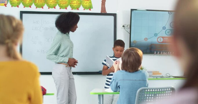 Little teen African American schoolboy standing in the middle of classroom and answering question while reading something from textbook in front of classmates and pretty teacher. Education concept