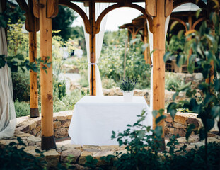 Garden gazebo with stone walling and wooden columns topped with architectural arches.In middle is table with white tablecloth and flower pot with plants.Garden vegetation in periphery is out of focus.