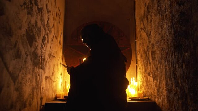 A man occultist in black robes stands with a candle and a knife in his hand