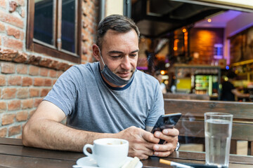 Mature caucasian man sitting in the restaurant or cafe for vaccinated guest using mobile phone while sitting by the table and wearing face mask on chin delta variant corona virus spread