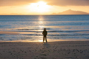 Naklejka premium Golden glow of sunrise across sea with silhouette of boy standing on beach on beach Waipu Cove