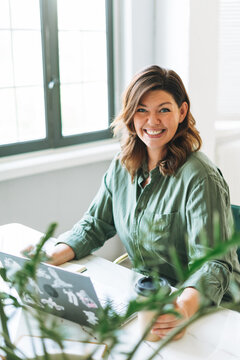 Young smiling brunette woman plus size working at laptop on table with house plant in bright modern office