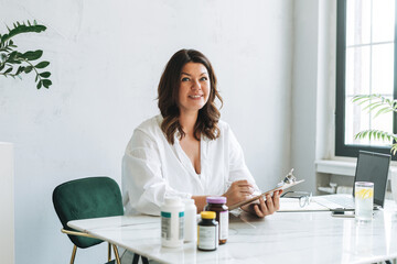 Young smiling brunette woman nutritionist plus size in white shirt working at laptop on table with house plant in the bright modern office