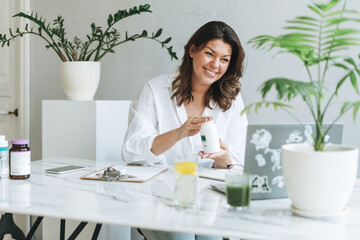 Young smiling brunette woman nutritionist plus size in white shirt working at laptop on table with house plant in the bright modern office. Doctor communicates with patient online