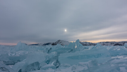 There is a block of turquoise hummocks on the surface of the frozen lake. Highlights on the edges. The setting sun in a cloudy sky. A mountain range in the distance. Baikal