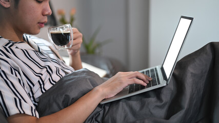 Casual man drinking hot coffee and using laptop computer on his bed in the morning.
