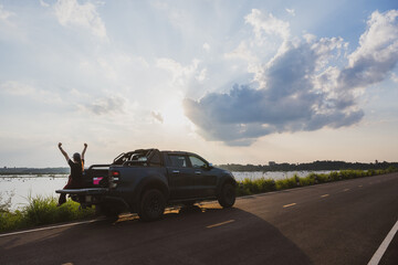 Fototapeta premium Asian female traveler camping in the back of a car with a background near a lake or river in vintage tones happy young woman traveling in summer country road girl sitting in the back of the car