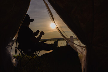 Young Asian traveler camping and making his own drip coffee relaxing in front of a camping tent in the evening against a lake or river background. Happy young Asian man goes on a summer trip.