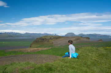 Naklejka na ściany i meble Person sitting on a grass enjoying view to the green mountains and valley, Iceland