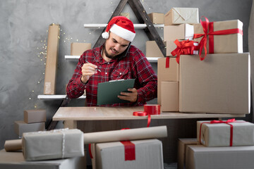 A salesman in a Santa Claus hat takes an order by cell phone while sitting in an office with boxes of gifts for Christmas. Shopping and delivery