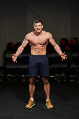 Fototapeta na wymiar a pumped up young man with a naked torso in a gym tensing muscles on his arms and chest with a cheerful smile