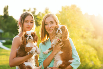 Horizontal authentic portrait of Breeder. Mother and daughter walking at sunset on summer day with pets. Puppies Cavalier King Charles Spaniel with owners outdoors.