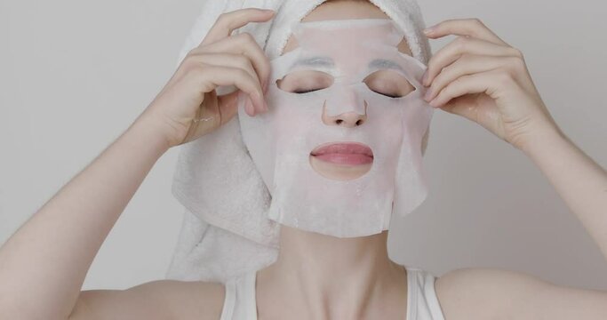 Anti-aging and hydrating spa treatments., sheet masks. Woman with white towel on her head applying hydrating sheet mask on her face. Skin care cosmetics. Isolated on gray background