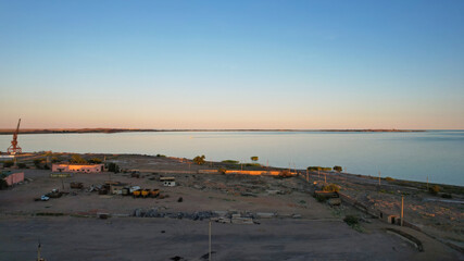 Abandoned territory on the beach of Lake Balkhash. The gradient of the sky at sunset. There is a crane and old fences, buildings. People are walking on the beach. Reeds grow in the water. Kazakhstan
