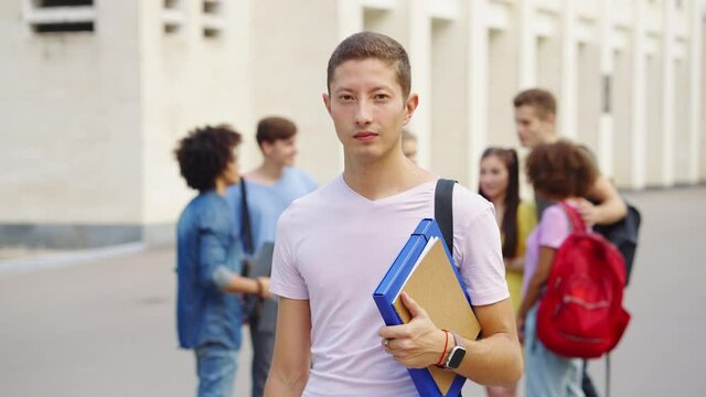 Student in casual wear with watch and red thread on wrist standing with file folder and exercise book near college and looking at camera, friends meeting on blurred background. Concept of education