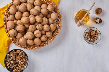 A wicker dish with walnuts stands on a white wooden table, next to a jar of honey and a bowl of peeled nuts.