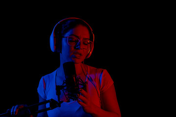 Caucasian woman in glasses and headphones sings into a microphone in neon light on a black background. An emotional girl is recording a song in a recording studio