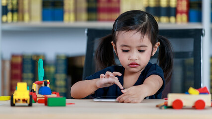 Small Asian pigtails hairstyle preschooler kindergarten girl sitting at table full of plastic wood truck toys smiling look at camera touching tablet computer in living room at home in front bookshelf