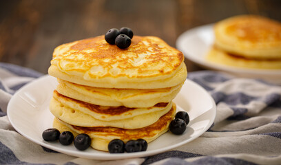 Stack of delicious pancakes sweet food with blueberries and blur dry leaves, plaid fabric in white plate on wooden background.