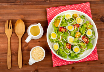 Big white bowl of lettuce, sprout salad with boil egg, and tomato mix with sesame dressing on red polka dots fabric above wooden table and small olive oil jar, sesame cup and utensil beside