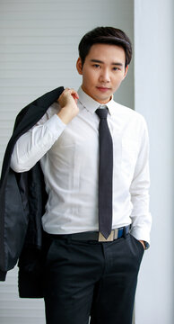 Asian men wearing white shirts, black tie, short hair, dark eyebrows smiling standing looking camera Hand holding suit across the back one hand put in pants pocket photo shooting in the white office.