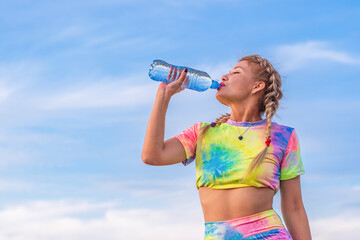 Young woman in a bright multicolored tracksuit drinks water from a transparent plastic bottle...