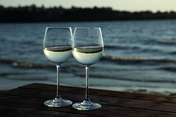 Two glasses of wine on wooden table near river at sunset. Space for text