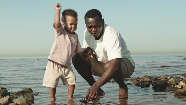 Slowmo shot of cute African-American toddler boy standing with his bare feet in water at beach together with dad, throwing pebble stones in water