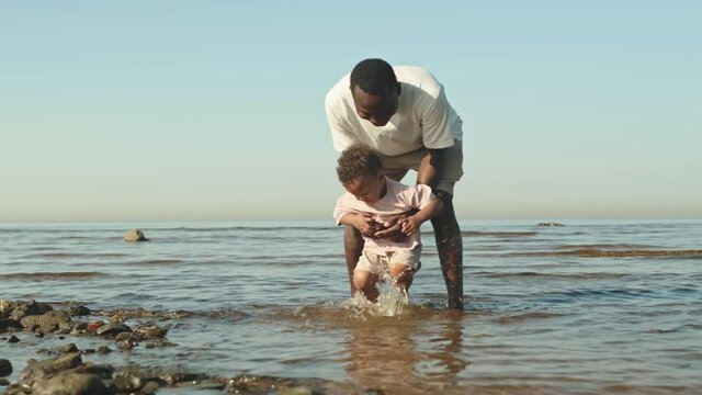 Slowmo shot of affectionate African-American father holding his playful toddler son on hands standing in water at beach. Boy having fun splashing water with his feet