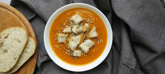 Orange pumpkin soup with croutons, sunflower seeds, chia seeds and sesame seeds in a white plate on a dark linen napkin. Two slices of bread on a wooden plate. Cooking template.