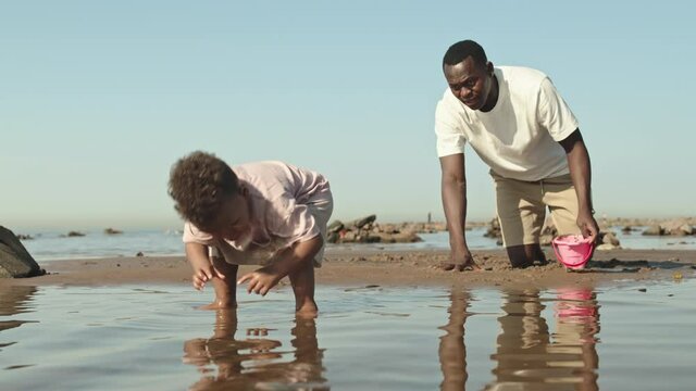 Slowmo shot of little African-American toddler boy playing in water and his father watching him sitting in sand at seashore spending summer day together