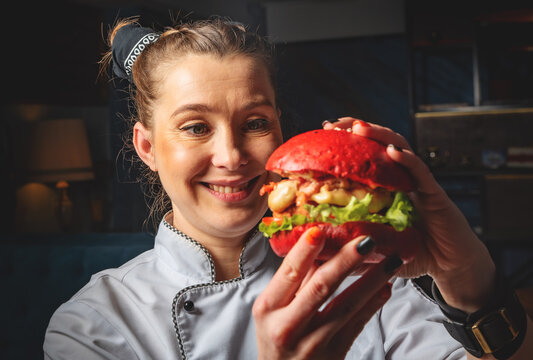 young woman chef with burger in hands on dark background in cafe