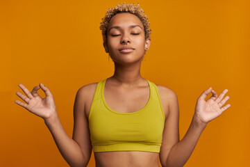 Portrait of relaxed, meditating African woman reaching zen and inner harmony, making Om sign with fingers, looking peaceful, feeling balanced enjoying spiritual practice isolated on orange wall - obrazy, fototapety, plakaty