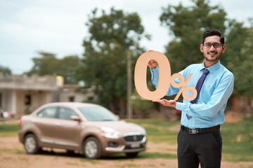 vehicle loan concept : Young indian banker or financier showing zero percent sign symbol on new vehicle