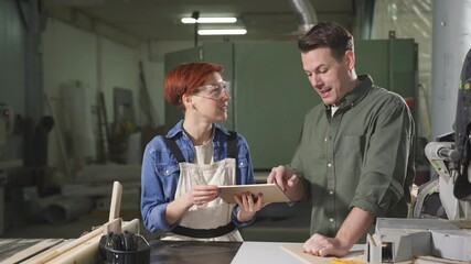 Young caucasian man and woman joiners using digital tablet while working in workshop, professional carpenters discussing working process, what to do, handmade and carpentry concept