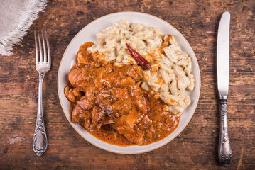 Portion of chicken paprikash with gnocchi or dumplings in a plate with a fork and knife on a wooden table, top view