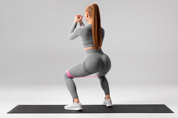 Athletic girl doing squats exercise for glute with resistance band on gray background. Fitness...